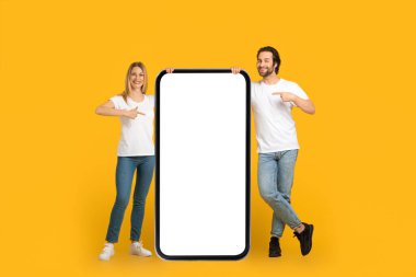 Tatmin olmuş milenyum Avrupalı beyaz tişörtlü çift boş ekranlı koca bir telefonu işaret ediyor.