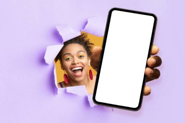 Smiling excited pretty young black woman with open mouth looks through hole in purple paper