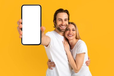 Beyaz tişörtlü mutlu beyaz erkek ve kadın sarılıyor, boş ekranlı akıllı telefonu gösteriyor.