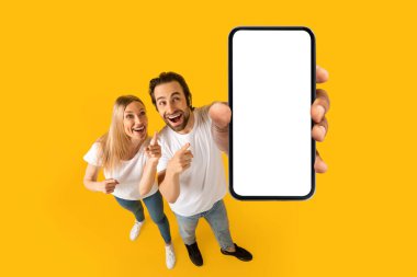 Glad surprised excited young european couple in white t-shirts show thumbs up at smartphone with blank screen