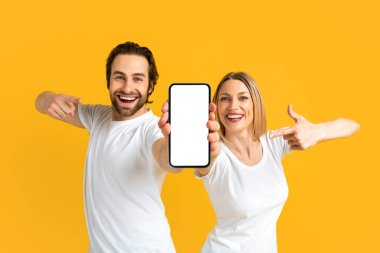 Glad excited young caucasian couple in white t-shirts pointing fingers at smartphone with empty screen