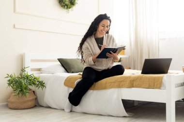 Yatakta oturan, bilgisayar kullanan ve not defterine yazan bir kadın.