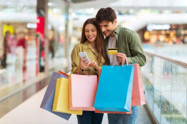 Couple Shopping With Cellphone And Credit Card In Modern Mall
