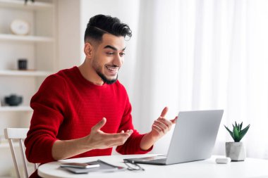 Gülümseyen, sakallı, genç, ortadoğulu bir erkek bilgisayara bakıyor ve iş yerinde el kol hareketi yapıyor.