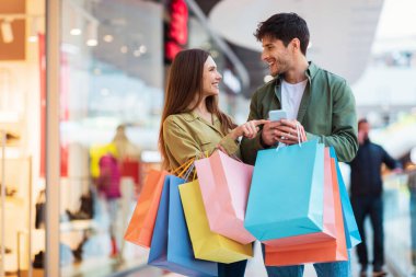 Cheerful Couple Shopping Using Phone Holding Shopper Bags In Hypermarket