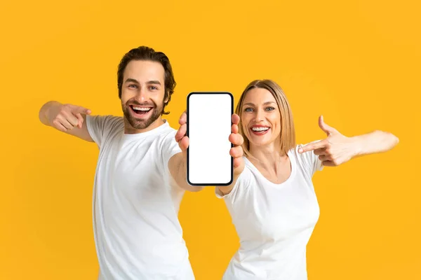 Feliz joven pareja caucásica emocionada en camisetas blancas apuntando con los dedos al teléfono inteligente con la pantalla vacía — Foto de Stock