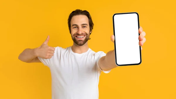 Glad young european man with stubble in white t-shirt show smartphone and thumb up with blank screens — Stockfoto