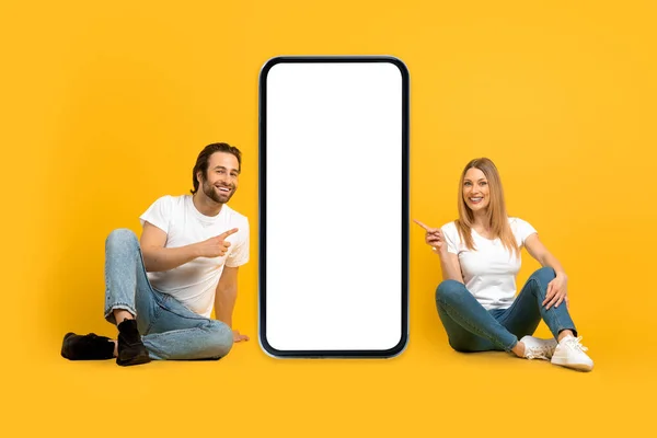Jovem mulher europeia feliz e marido com restolho sentar no chão, apontar os dedos para o smartphone grande com tela em branco — Fotografia de Stock