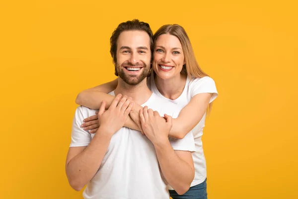 Cheerful millennial european female hugging from behind male with stubble in white t-shirts looking at camera — Fotografia de Stock