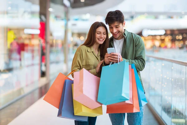 Cônjuges felizes fazendo compras e usando celular em pé no shopping — Fotografia de Stock