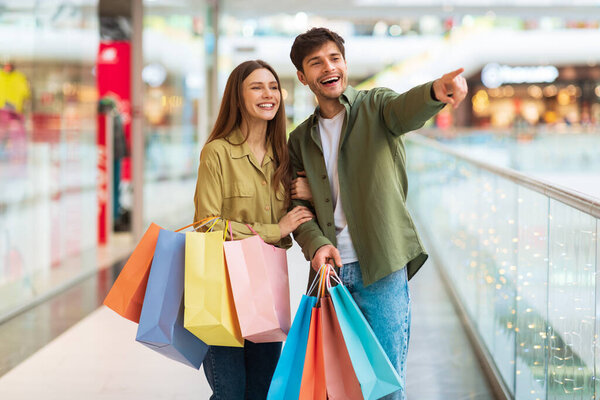 Happy Spouses Holding Shopper Bags Pointing Finger Shopping In Hypermarket