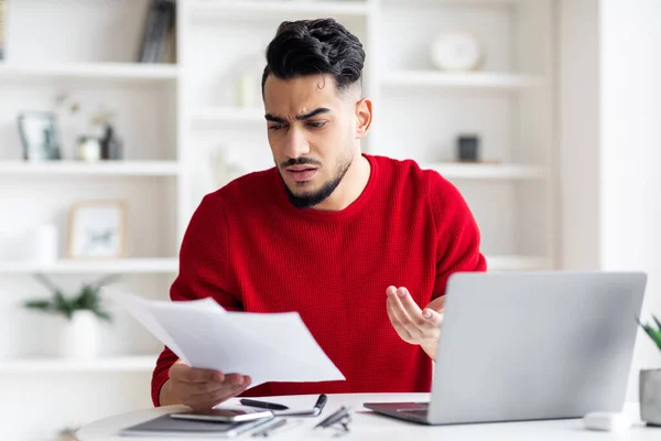 Sad worried young arab man with beard works with documents and cant find at workplace with laptop — Stockfoto