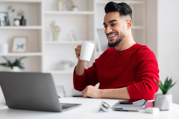 Glücklicher junger islamischer Mann mit Bart trinkt Tasse Tee am Arbeitsplatz mit Computer im Home Office Interieur — Stockfoto