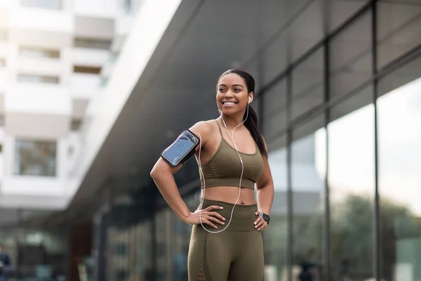 Joven mujer negra en ropa deportiva escuchando música, usando smartphone y rastreador de fitness durante el jogging en la calle de la ciudad —  Fotos de Stock