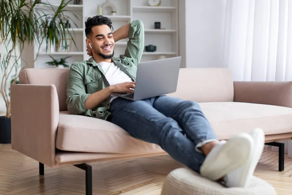 Cheerful millennial arab man with beard sits on sofa in wireless headphones and typing on laptop — Φωτογραφία Αρχείου