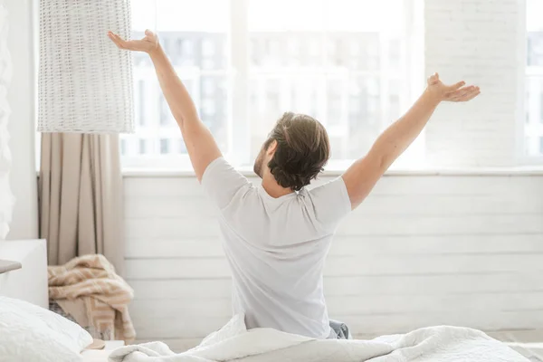 Happy millennial caucasian guy wake up and stretching body, sitting on soft bed on window background — Stock Photo, Image