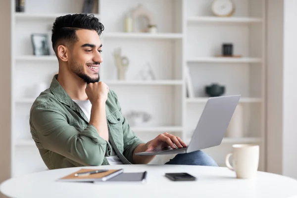 Feliz joven árabe hombre con barba mira a la computadora portátil, tener chat en las redes sociales en el interior de la oficina en casa — Foto de Stock