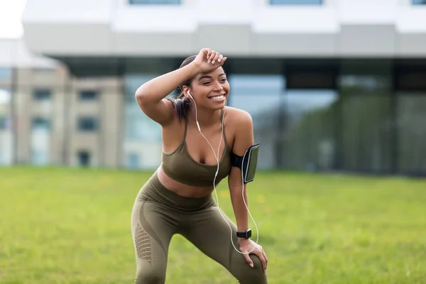 Felice stanca giovane donna afroamericana con il cellulare che ascolta musica durante il jogging mattutino, asciugando il sudore al parco — Foto Stock