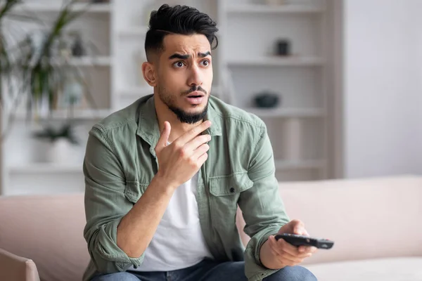 Shocked frightened young arab man with beard with remote control watches news on TV sits on sofa — Stockfoto