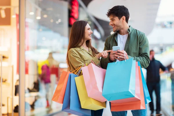 Cheerful Couple Shopping Using Phone Holding Shopper Bags In Hypermarket — Fotografia de Stock