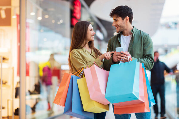 Cheerful Couple Shopping Using Phone Holding Shopper Bags In Hypermarket