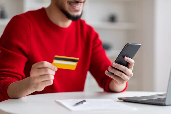 Pria arab muda tersenyum dengan jenggot menunjukkan kartu kredit dan mengetik di smartphone di interior kantor pusat — Stok Foto