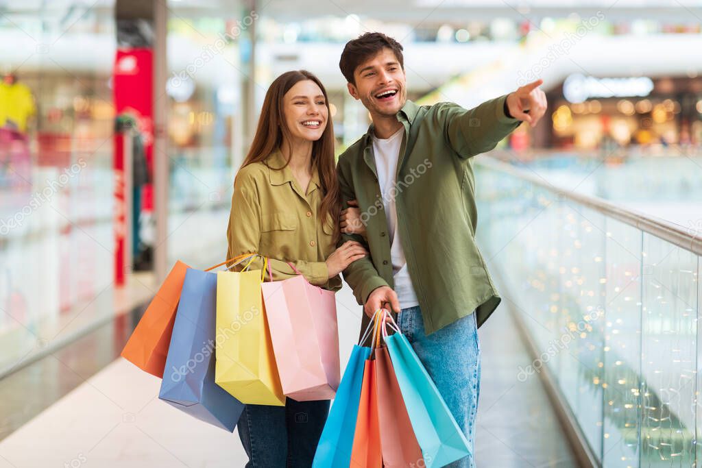 Happy Spouses Holding Shopper Bags Pointing Finger Shopping In Hypermarket
