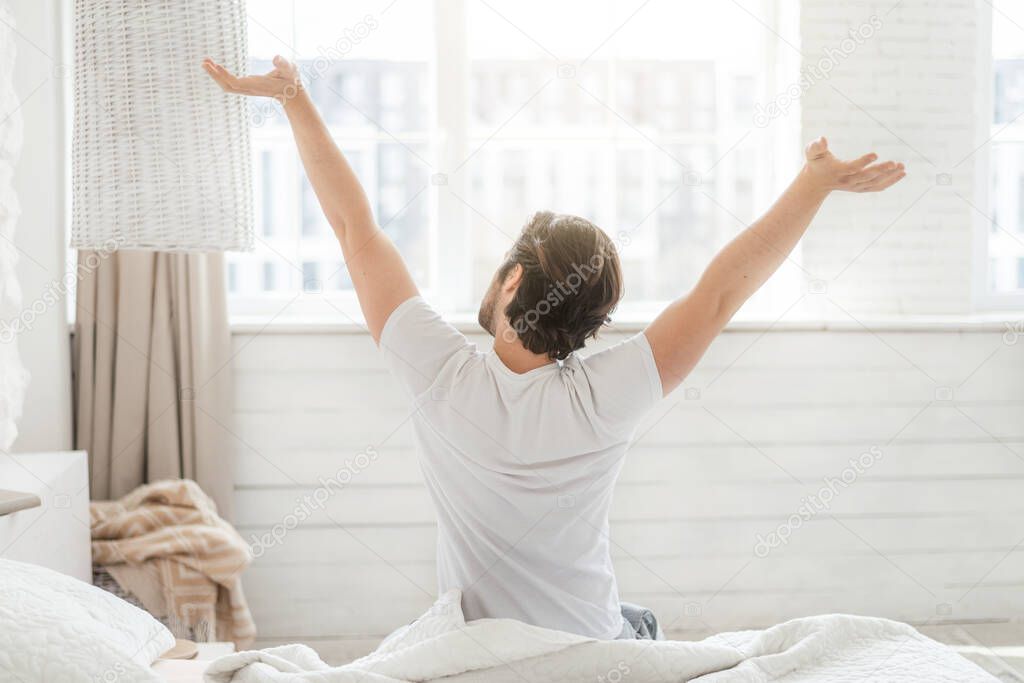 Happy millennial caucasian guy wake up and stretching body, sitting on soft bed on window background