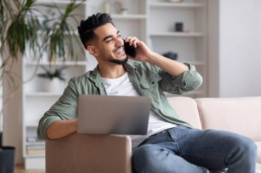 Cheerful arab man with beard sits on sofa with laptop, talk by phone, looks at empty space in living room