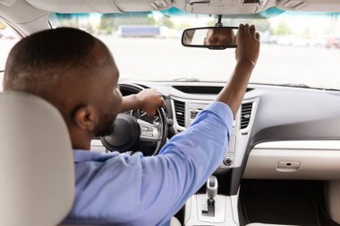 Gülümseyen siyah adam şehirde yeni araba kullanıyor, aynayı ayarlıyor.