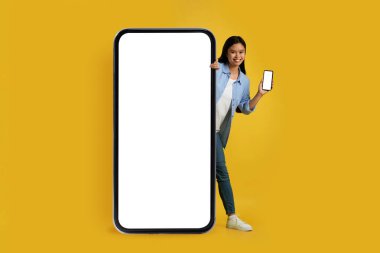 Smiling funny young asian female student in casual with smartphone peeking out from huge phone with empty screen
