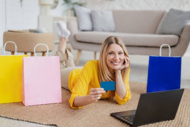 Dizüstü bilgisayarı ve kredi kartı olan, memnun, genç, sarışın Avrupalı bayan yerde bir sürü paketle yatıyor.