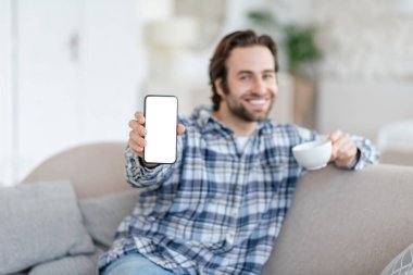 Smiling young caucasian man with stubble sits on sofa with coffee cup, shows phone with blank screen