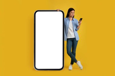 Akıllı telefona sıradan bir bakışla bakan mutlu genç Japon kadın boş ekranlı dev bir telefonun yanında duruyor.