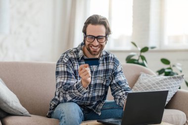 Happy young caucasian man with stubble in glasses looks at credit card and sits on sofa with laptop
