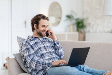 Bin yıllık neşeli, sakallı beyaz erkek kanepeye oturur, dizüstü bilgisayarda yazar ve telefonla arar.