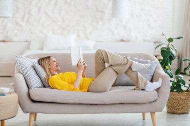 Sarı elbiseli mutlu, genç, sarışın Avrupalı kadın kanepeye uzanıp kitap okuyor.