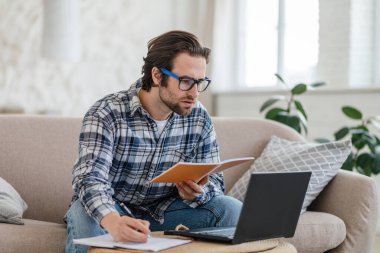 Bin yıllık beyaz tenli, gözlüklü bir erkek kanepeye oturup dizüstü bilgisayar ve kitapla notlar alıyor.