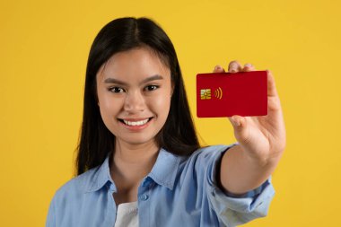 Sarı arka planda izole edilmiş, sıradan bir şekilde kredi kartı gösteren neşeli, genç Koreli bir kadın.