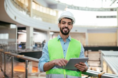 Sakallı, sakallı, tabletli koruyucu üniformalı gülümseyen genç Arap adam.