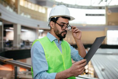 Ciddi çekici genç Arap mühendis kasklı sakallı koruyucu üniformalı gözlüğünü çıkarıyor.