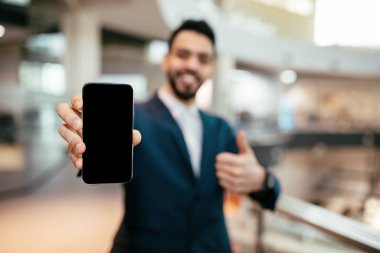 Smiling young arab man with beard in suit shows smartphone with blank screen and thumb up in office