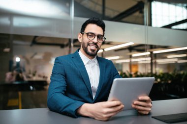 Gözlüklü ve takım elbiseli neşeli, çekici, orta doğulu erkek ofiste tabletle masada oturuyor.