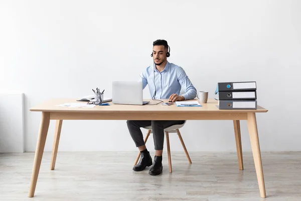 Uomo arabo che utilizza il computer portatile indossando auricolare seduto alla scrivania — Foto Stock