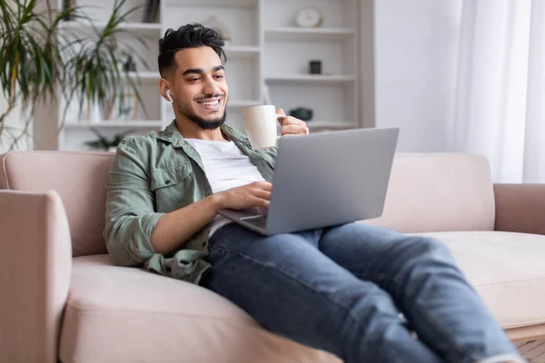 Glad millennial islamic guy with beard sits on sofa in wireless headphones, watch laptop, enjoy coffee and rest — Stockfoto