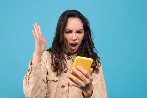 Sad shocked young european brunette woman with open mouth looks at smartphone isolated on blue background — Stok fotoğraf