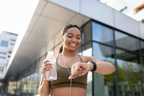 Donna nera che guarda smartwatch, monitoraggio attività di fitness, ascoltare musica, acqua potabile durante l'allenamento all'aperto — Foto Stock