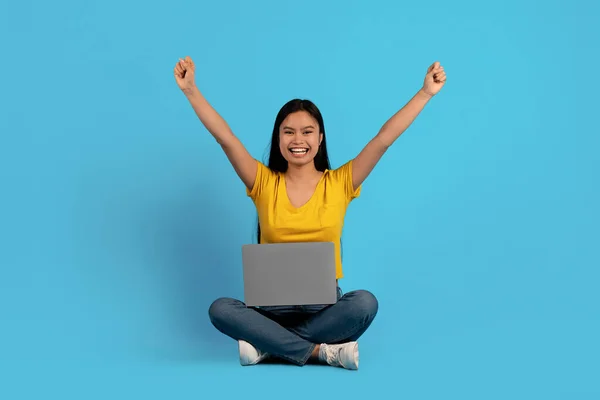 Glad young chinese lady in casual sits on floor with laptop and raises hands up rejoices in victory —  Fotos de Stock