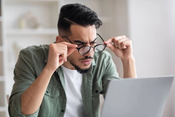 Здивований серйозний молодий арабський чоловік з бородою знімає окуляри дивиться на ноутбук в інтер'єрі домашнього офісу — стокове фото