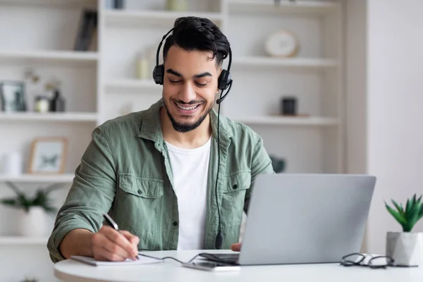 Felice giovane arabo con la barba in cuffia funziona sul computer portatile, guardare webinar, prendere appunti — Foto Stock